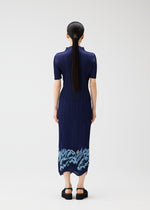 A woman with long, dark hair in a low ponytail faces away, wearing the PLEATS PLEASE ISSEY MIYAKE I SPY! navy dress with a pleated, embroidered skirt featuring light blue floral details, paired with black platform sandals.