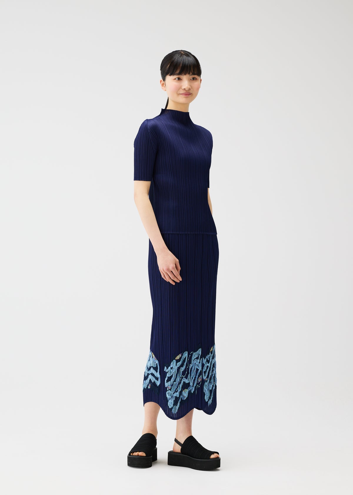 A woman with short dark hair stands against a white background, wearing the navy I SPY! ankle-length embroidered skirt by PLEATS PLEASE ISSEY MIYAKE, featuring light blue patterns near the hem and paired with black platform sandals. view 8 of 10