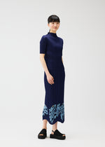 A woman with short dark hair stands against a white background, wearing the navy I SPY! ankle-length embroidered skirt by PLEATS PLEASE ISSEY MIYAKE, featuring light blue patterns near the hem and paired with black platform sandals.