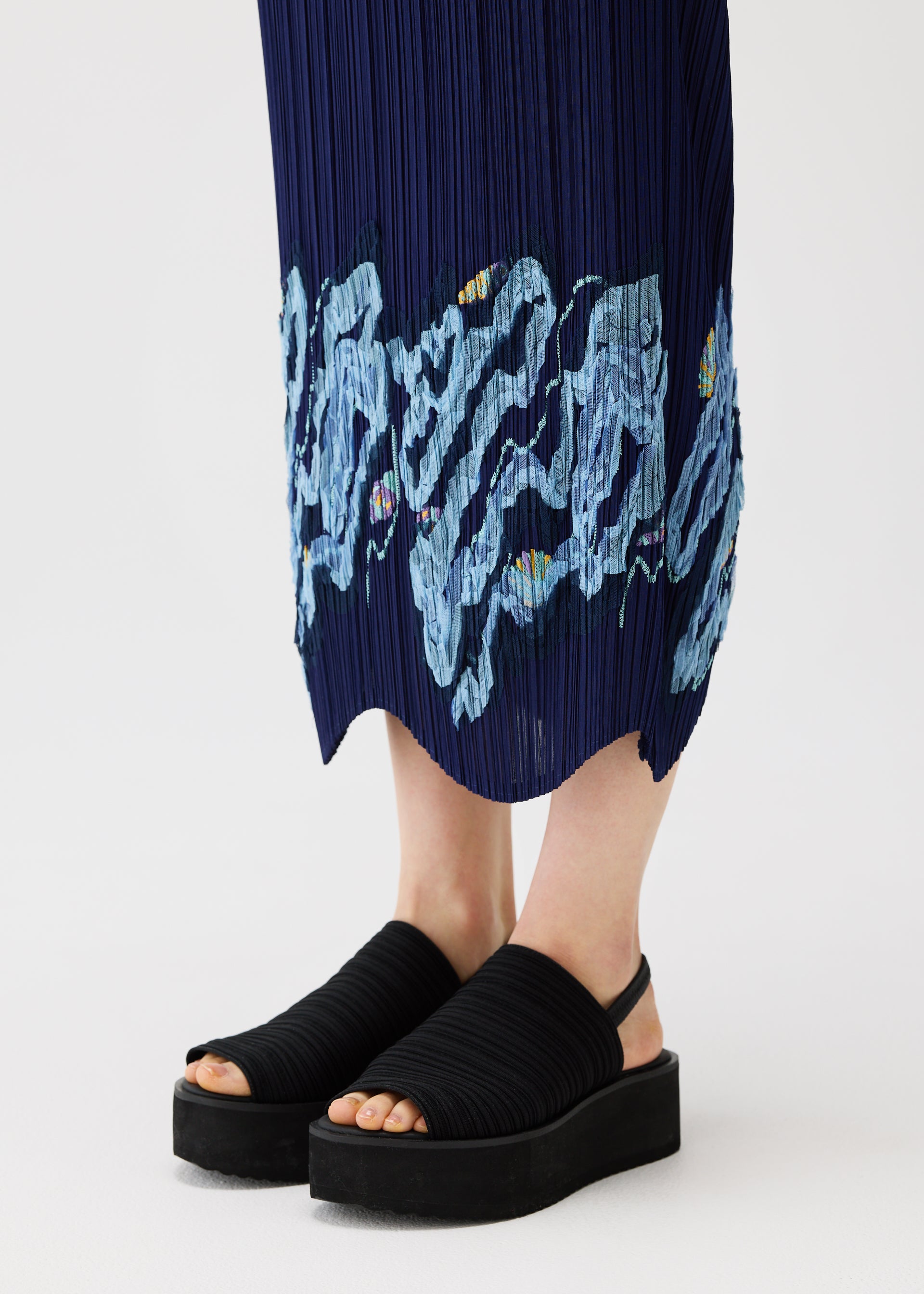 Zoomed view A person wearing black platform sandals stands on a white surface, dressed in the PLEATS PLEASE ISSEY MIYAKE I SPY! navy pleated skirt featuring wavy light blue embroidery near the hem.