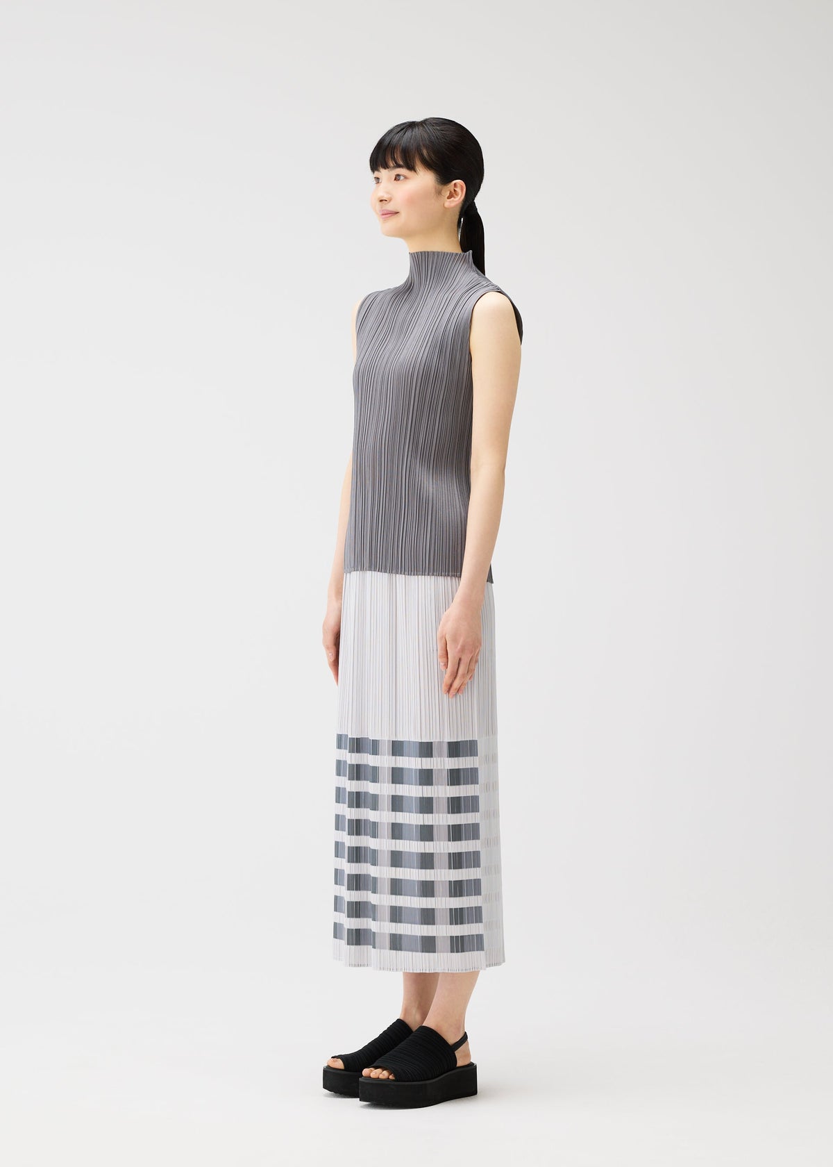 A woman stands sideways against a plain white background, wearing the PLEATS PLEASE ISSEY MIYAKE BUMPY BASKET sleeveless gray and white long dress with pleated texture, striped hem, and black platform sandals. view 6 of 8
