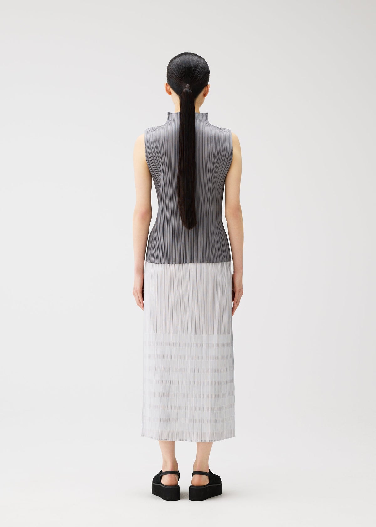 A person with long, straight black hair in a low ponytail stands facing away, wearing the PLEATS PLEASE ISSEY MIYAKE BUMPY BASKET gray-to-white gradient skirt with pockets and black platform sandals against a plain white background. view 7 of 8