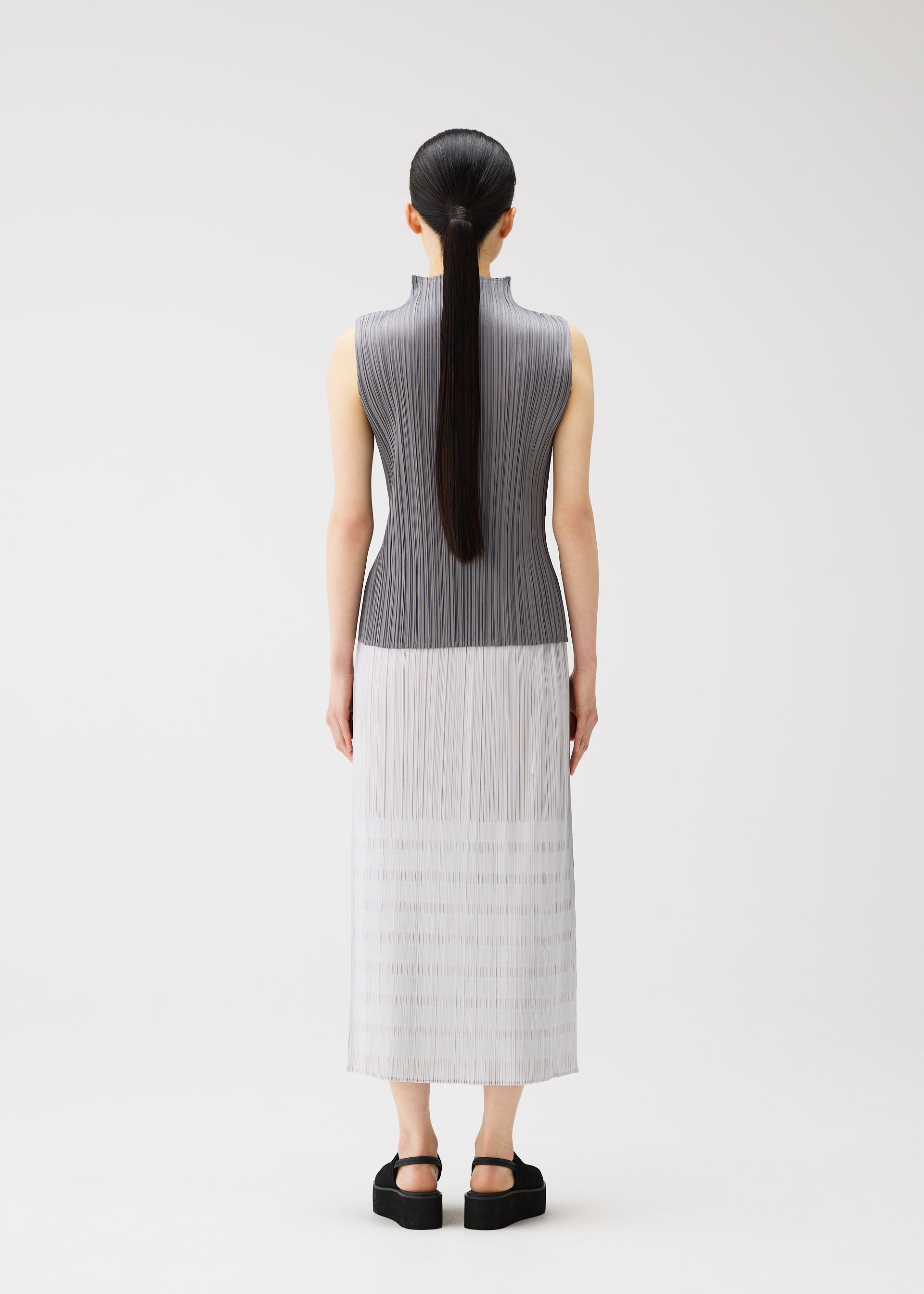Zoomed view A person with long, straight black hair in a low ponytail stands facing away, wearing the PLEATS PLEASE ISSEY MIYAKE BUMPY BASKET gray-to-white gradient skirt with pockets and black platform sandals against a plain white background.