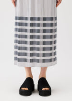 A person models the PLEATS PLEASE ISSEY MIYAKE BUMPY BASKET skirt, featuring long pleats with gray and white horizontal stripes and subtle pockets, paired with black platform slide sandals. Only the lower body is shown against a white background.