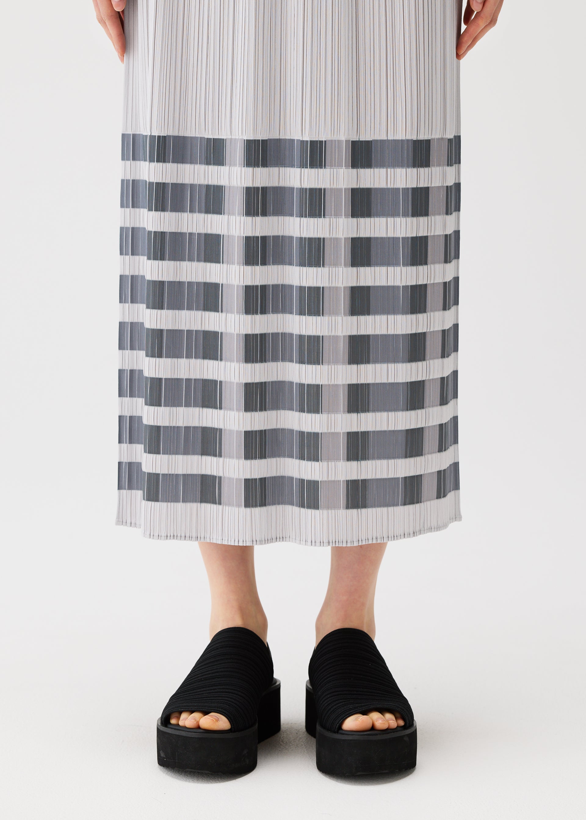 Zoomed view A person models the PLEATS PLEASE ISSEY MIYAKE BUMPY BASKET skirt, featuring long pleats with gray and white horizontal stripes and subtle pockets, paired with black platform slide sandals. Only the lower body is shown against a white background.