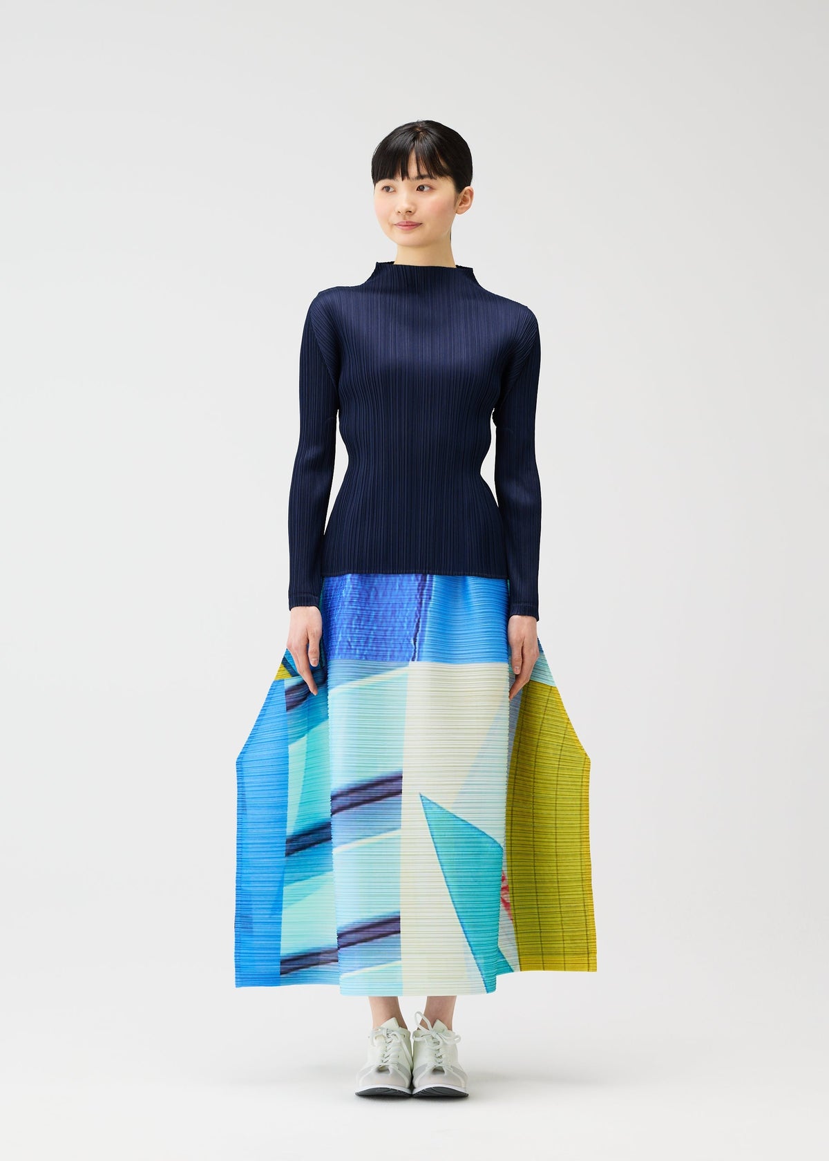 A woman poses against a white background wearing the CITY COLLAGE pleated skirt by PLEATS PLEASE ISSEY MIYAKE, featuring blue, aqua, and yellow townscape motifs, paired with a fitted navy top and white sneakers. view 6 of 9
