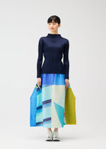 A woman poses against a white background wearing the CITY COLLAGE pleated skirt by PLEATS PLEASE ISSEY MIYAKE, featuring blue, aqua, and yellow townscape motifs, paired with a fitted navy top and white sneakers.