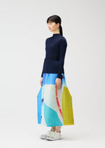 A woman in profile wears the CITY COLLAGE pleated skirt by PLEATS PLEASE ISSEY MIYAKE, featuring a unique townscape print, paired with a navy ribbed top and white sneakers against a plain white background.
