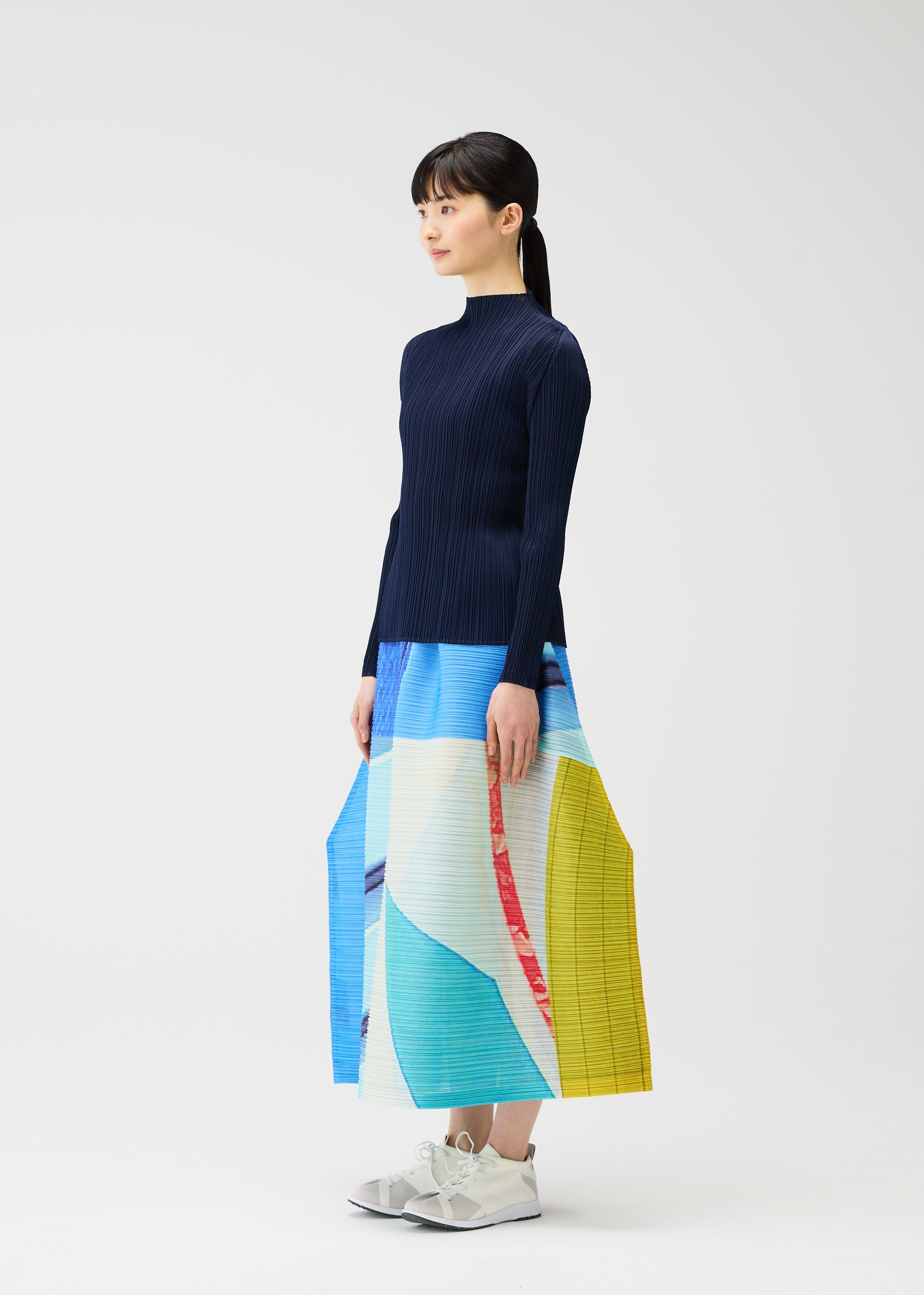 Zoomed view A woman in profile wears the CITY COLLAGE pleated skirt by PLEATS PLEASE ISSEY MIYAKE, featuring a unique townscape print, paired with a navy ribbed top and white sneakers against a plain white background.