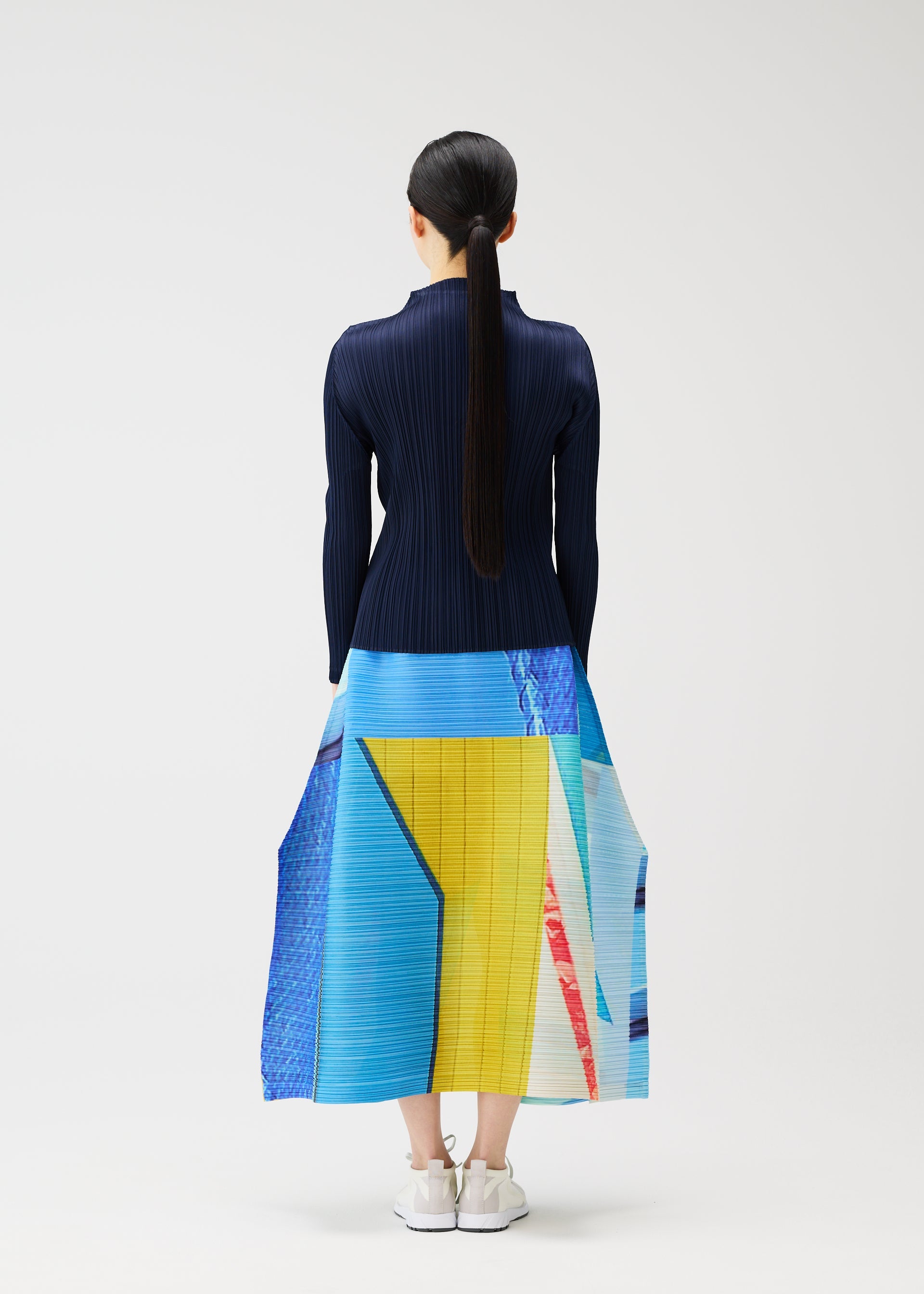 Zoomed view A woman with long dark hair in a ponytail faces away, wearing PLEATS PLEASE ISSEY MIYAKE's CITY COLLAGE navy top and pleated skirt featuring blue, yellow, and red townscape patterns, paired with white sneakers against a plain background.