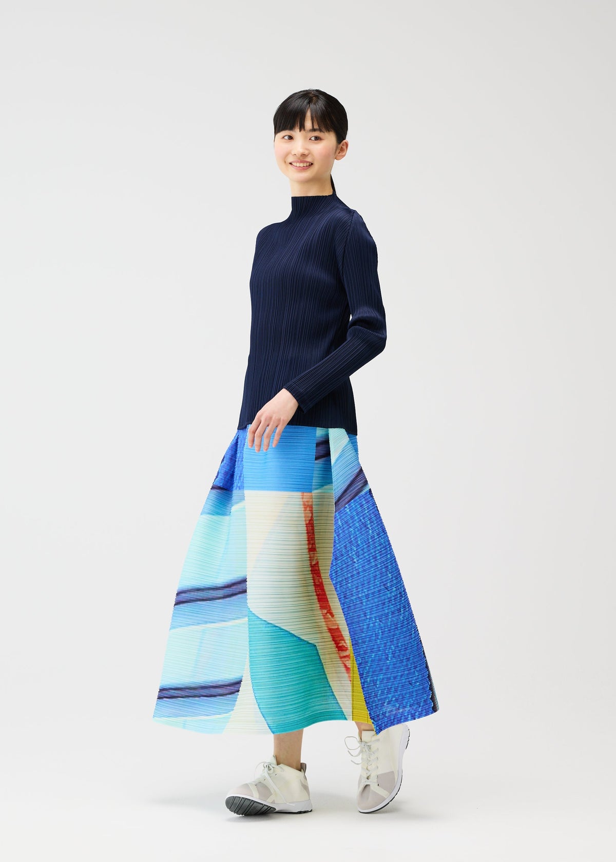 A person stands against a white background, smiling in the PLEATS PLEASE ISSEY MIYAKE CITY COLLAGE long, colorful pleated skirt with a unique townscape design, paired with a navy long-sleeve top and white sneakers. view 9 of 9
