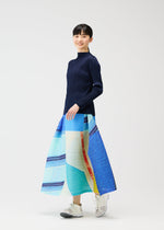 A person stands against a white background, smiling in the PLEATS PLEASE ISSEY MIYAKE CITY COLLAGE long, colorful pleated skirt with a unique townscape design, paired with a navy long-sleeve top and white sneakers.