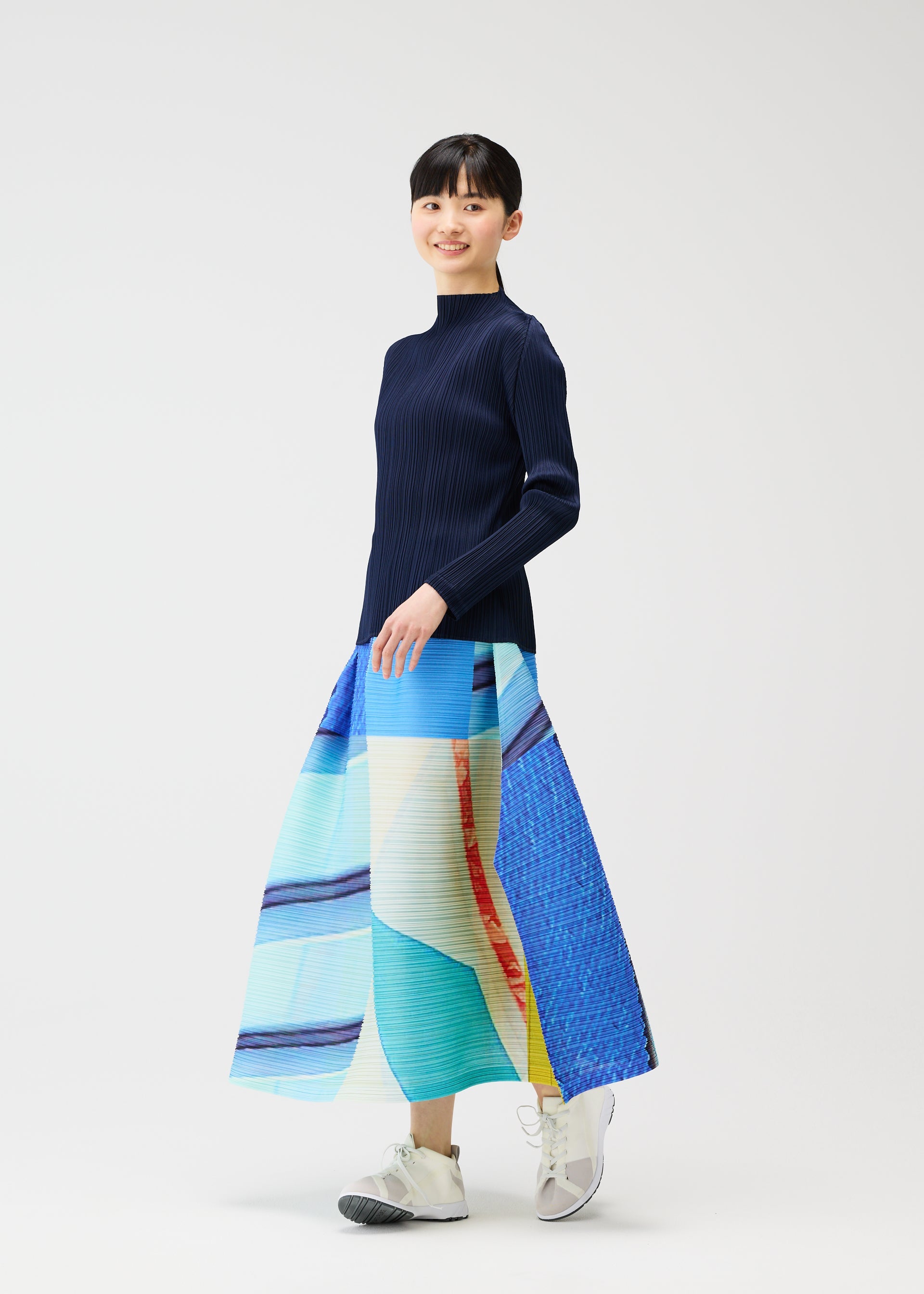 Zoomed view A person stands against a white background, smiling in the PLEATS PLEASE ISSEY MIYAKE CITY COLLAGE long, colorful pleated skirt with a unique townscape design, paired with a navy long-sleeve top and white sneakers.