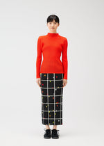 A woman faces forward in PLEATS PLEASE ISSEY MIYAKE's CROSSING DOTS—a bright red ribbed turtleneck sweater and a black skirt with a white grid and colorful dots—paired with black shoes, set against a plain backdrop.