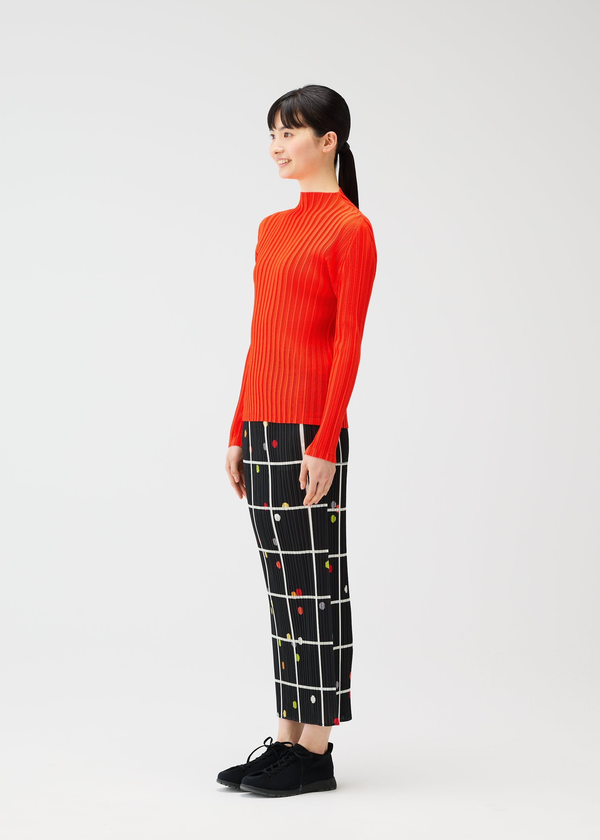 Zoomed view A woman stands sideways wearing the CROSSING DOTS skirt by PLEATS PLEASE ISSEY MIYAKE, paired with a bright orange ribbed sweater and black shoes, against a plain white background.