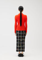 A woman with long, straight black hair in a ponytail faces away, wearing the CROSSING DOTS skirt by PLEATS PLEASE ISSEY MIYAKE—a black ankle-length plaid dot skirt with a white grid and colorful dots—paired with a bright red ribbed top and black shoes.