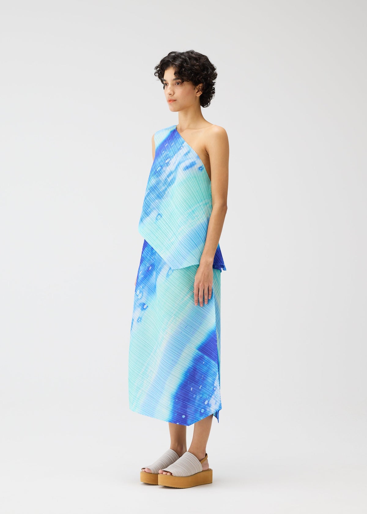 A person wears the SEA BUBBLE one-shoulder top and matching diagonal pleated skirt by PLEATS PLEASE ISSEY MIYAKE, featuring a blue and aqua abstract pattern, styled with platform sandals against a plain white background. view 5 of 7