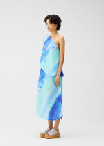 A person wears the SEA BUBBLE one-shoulder top and matching diagonal pleated skirt by PLEATS PLEASE ISSEY MIYAKE, featuring a blue and aqua abstract pattern, styled with platform sandals against a plain white background.