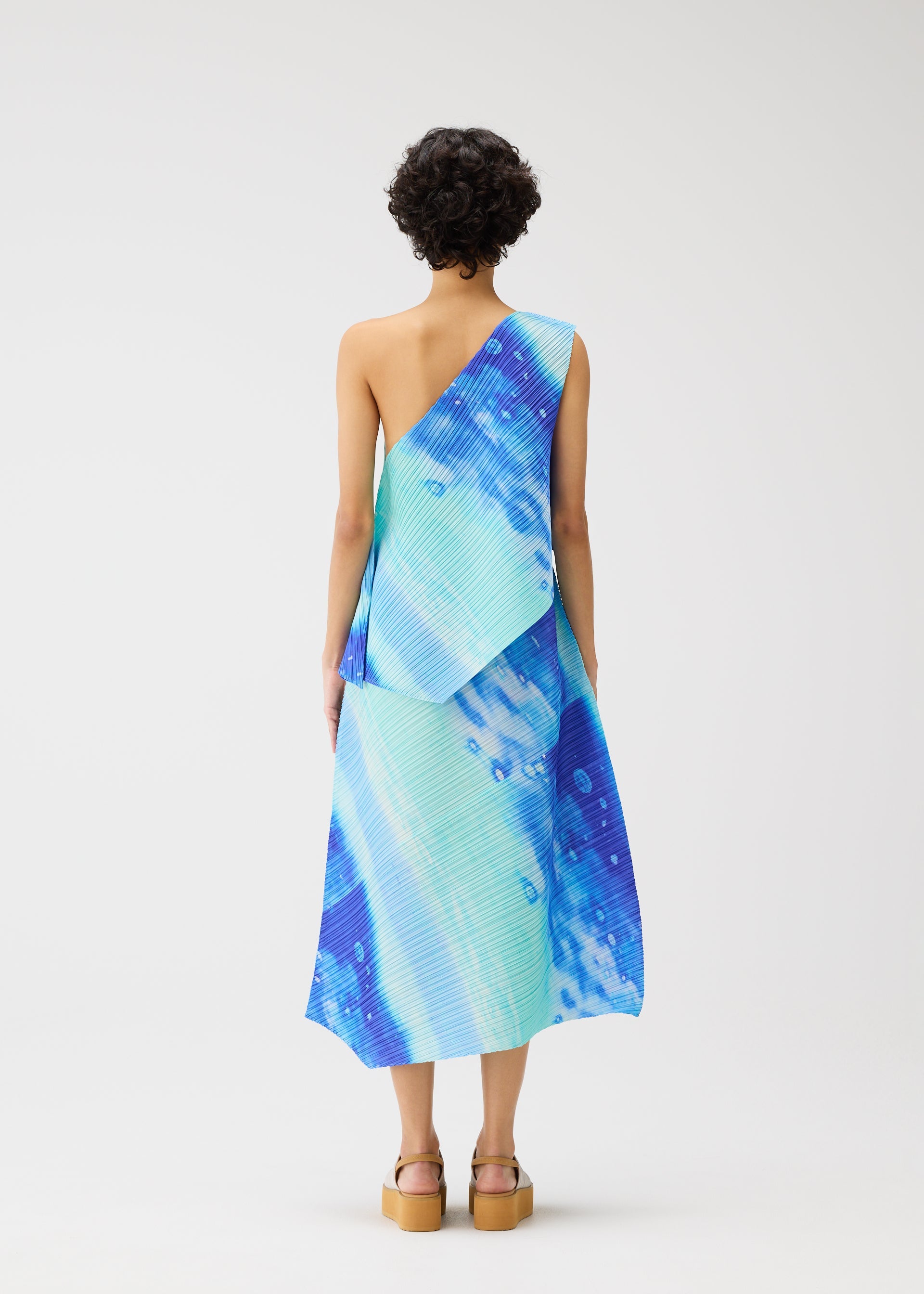 Zoomed view A person with short curly hair is seen from the back wearing the SEA BUBBLE one-shoulder dress by PLEATS PLEASE ISSEY MIYAKE in blue and turquoise, styled with tan platform sandals against a plain white background.