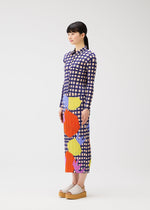 A woman in profile stands against a white background wearing the MARKET long-sleeve, ankle-length skirt dress with pockets by PLEATS PLEASE ISSEY MIYAKE, featuring a vegetable print of large colorful shapes, paired with beige platform sandals.