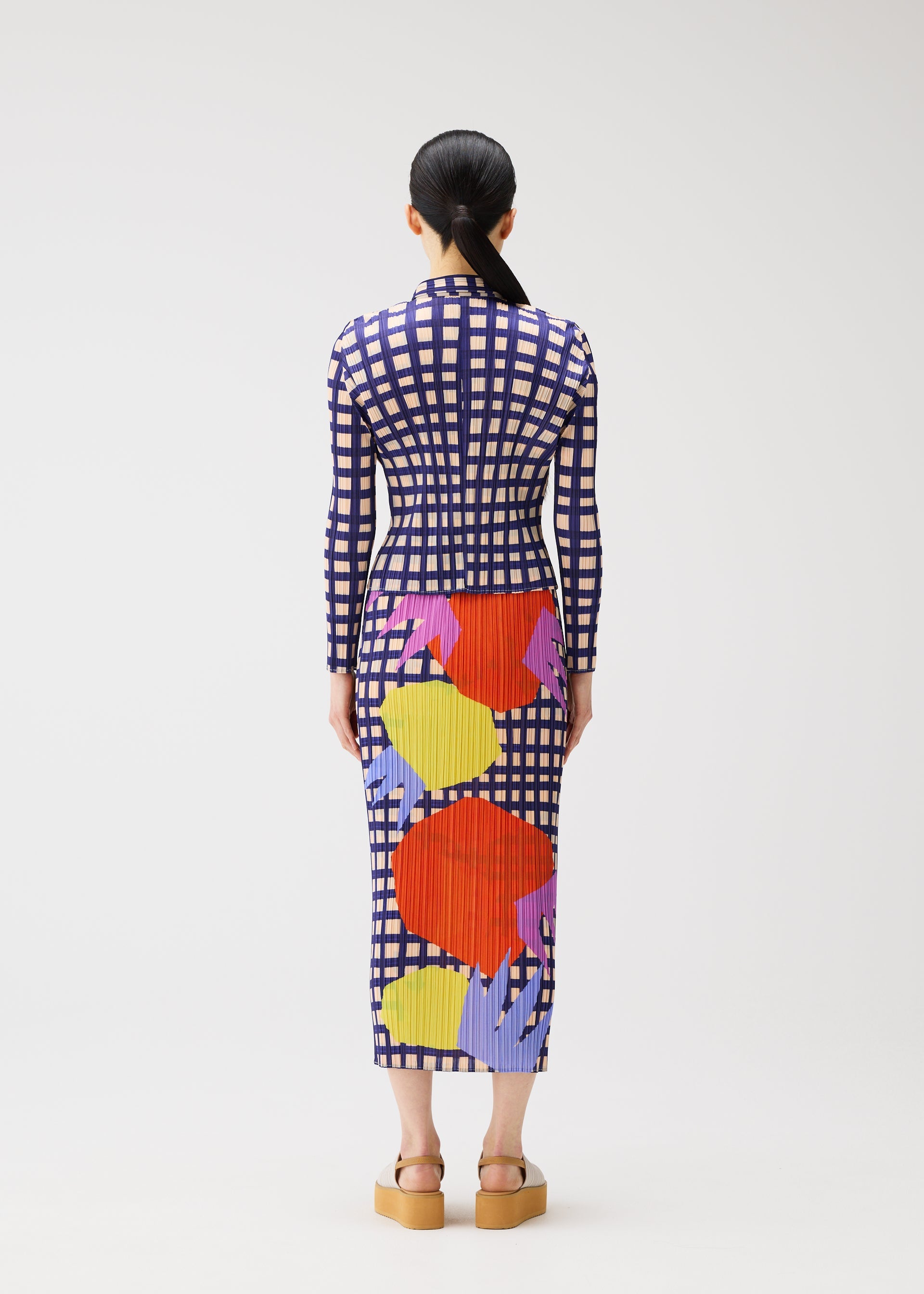 Zoomed view A person with long dark hair in a low ponytail is seen from behind wearing the MARKET dress by PLEATS PLEASE ISSEY MIYAKE, featuring a navy and white grid, colorful abstract shapes, pockets, and tan platform sandals.