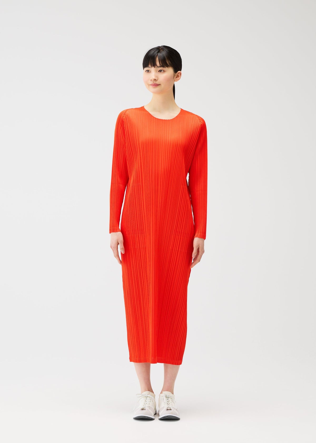 A woman with straight black hair and bangs wears a bright orange, long-sleeved MONTHLY COLORS : FEBRUARY dress by PLEATS PLEASE ISSEY MIYAKE and white sneakers, standing against a plain white background and smiling softly as she looks to the side. view 7 of 9