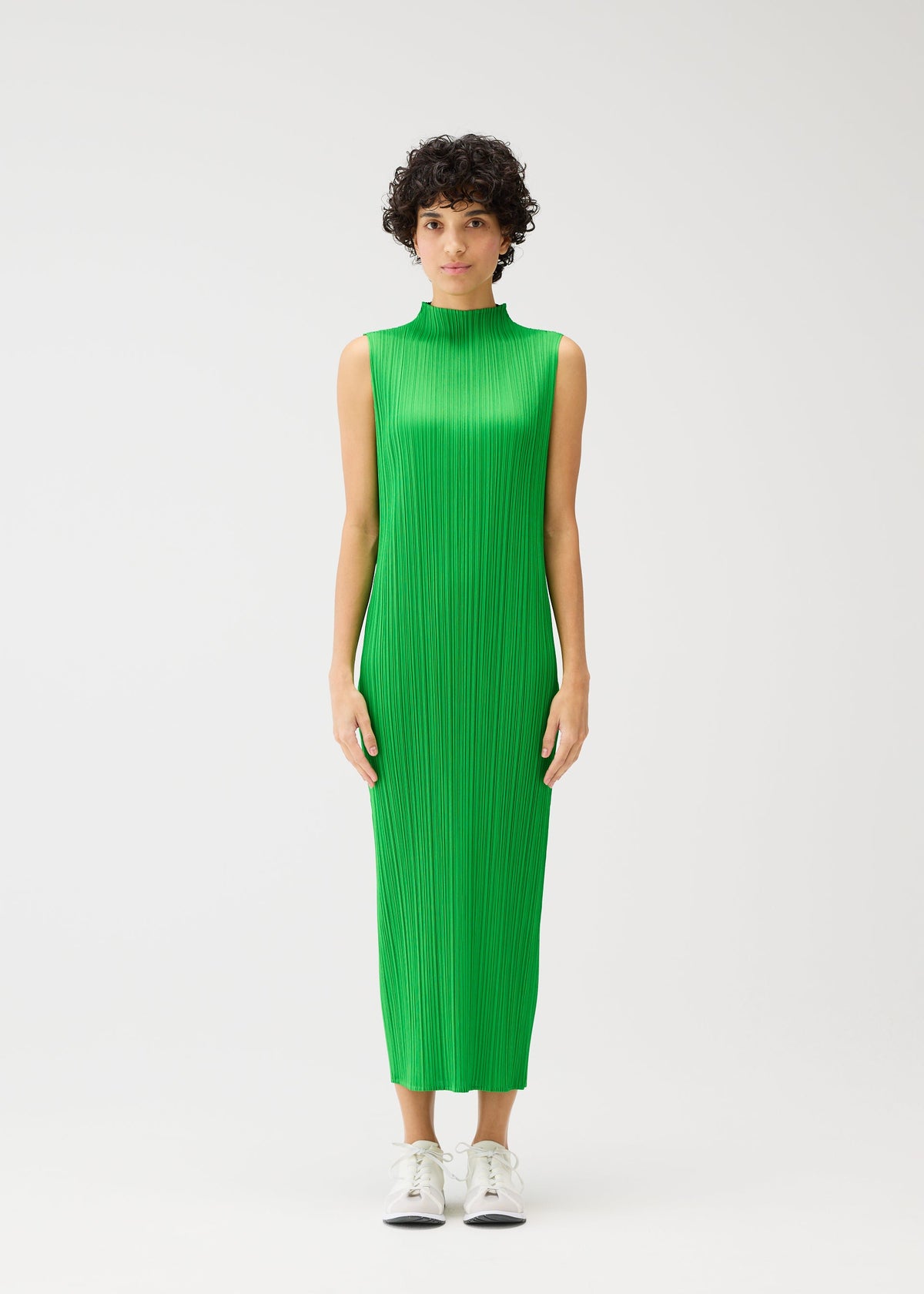 A person with short curly hair wears the MONTHLY COLORS : APRIL high-neck sleeveless dress by PLEATS PLEASE ISSEY MIYAKE in light green with vertical pleats, paired with white sneakers, standing against a plain white background. view 7 of 9
