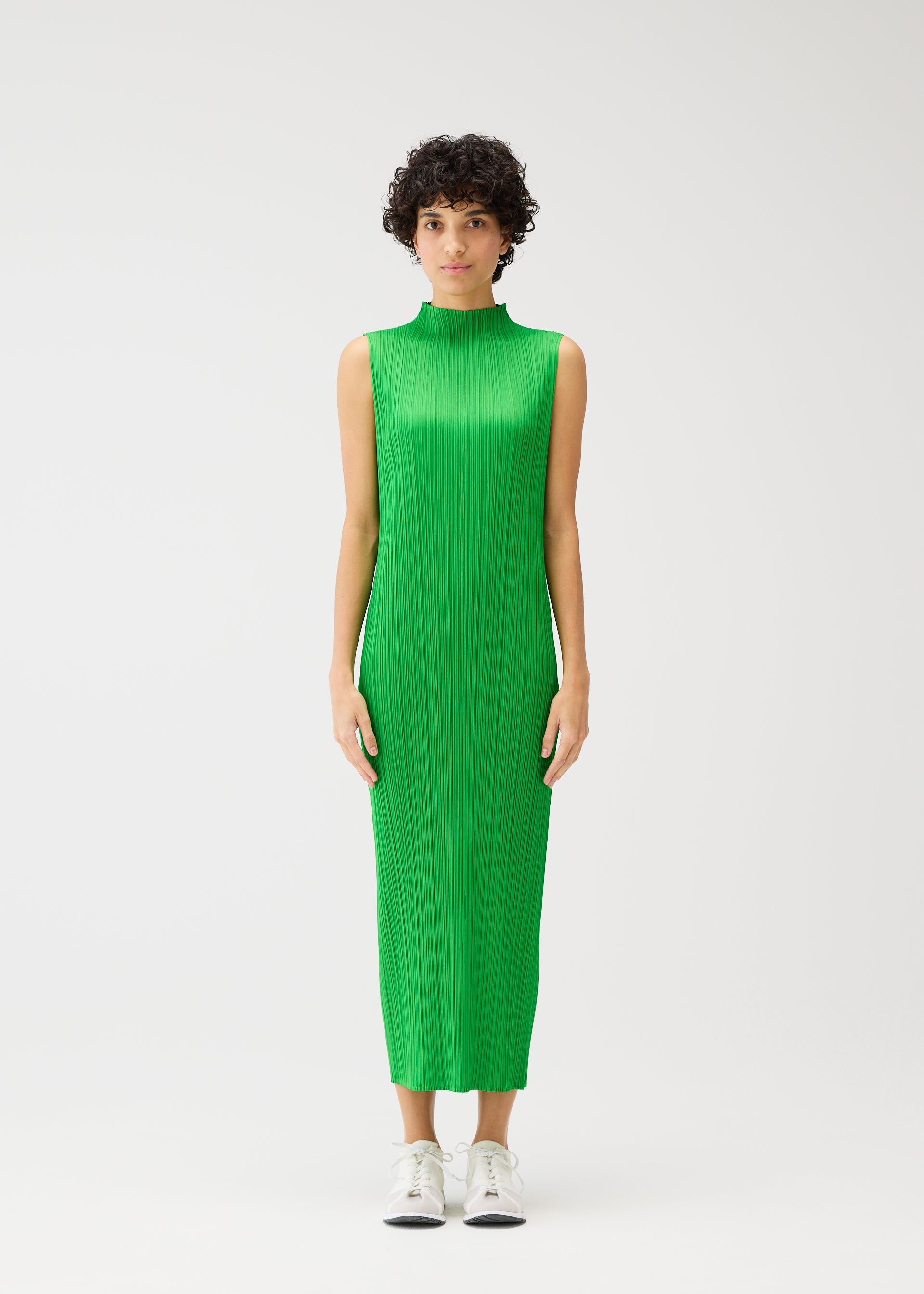 Zoomed view A person with short curly hair wears the MONTHLY COLORS : APRIL high-neck sleeveless dress by PLEATS PLEASE ISSEY MIYAKE in light green with vertical pleats, paired with white sneakers, standing against a plain white background.