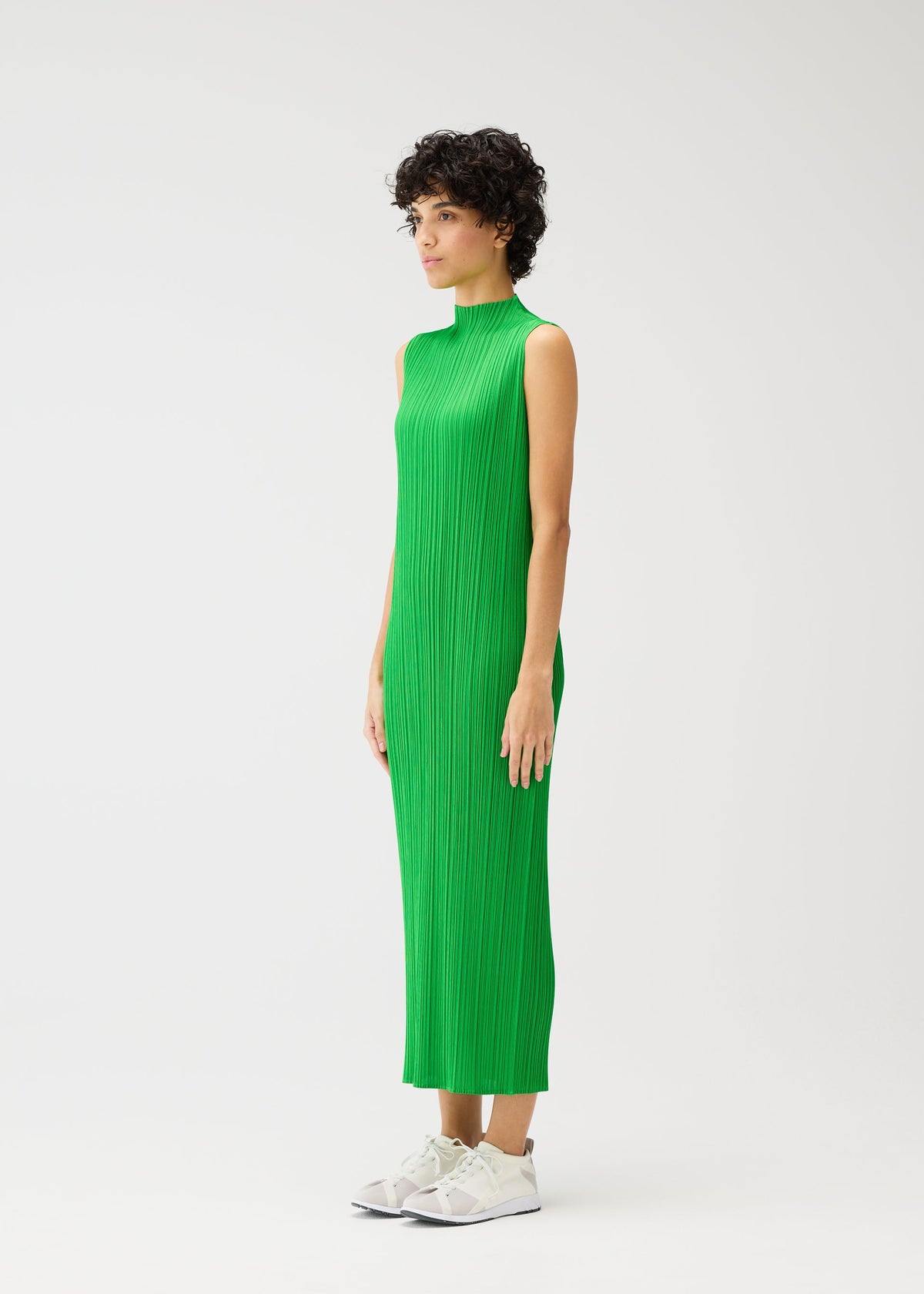 A person with short curly hair stands sideways against a white background, wearing PLEATS PLEASE ISSEY MIYAKE's MONTHLY COLORS : APRIL—an ankle-length, high-neck sleeveless bright green dress—and white sneakers. view 8 of 9
