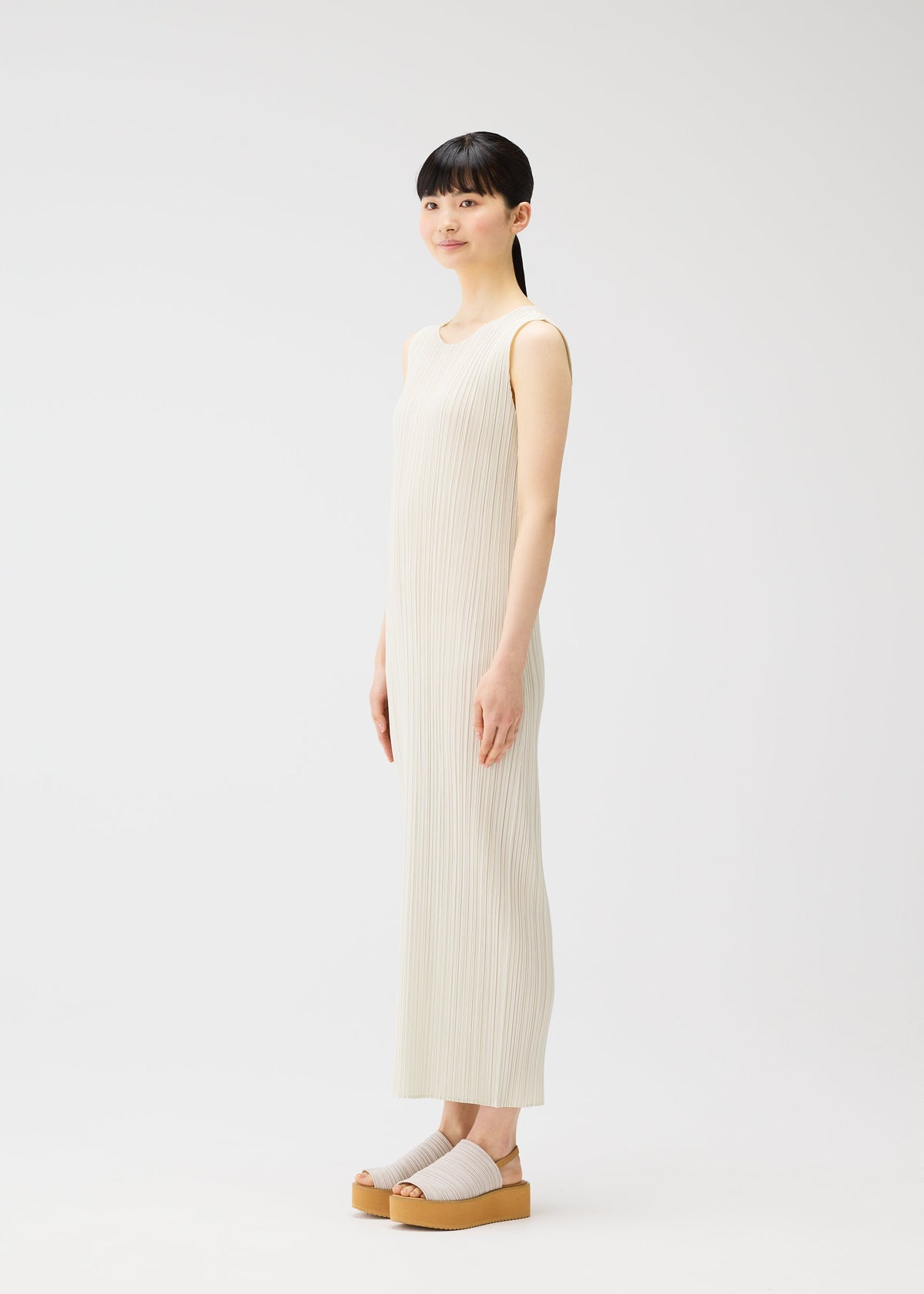 A woman with straight black hair and bangs stands sideways in a long, pleated dress from the MONTHLY COLORS : MAY collection by PLEATS PLEASE ISSEY MIYAKE, paired with beige platform sandals against a plain white background. view 8 of 9