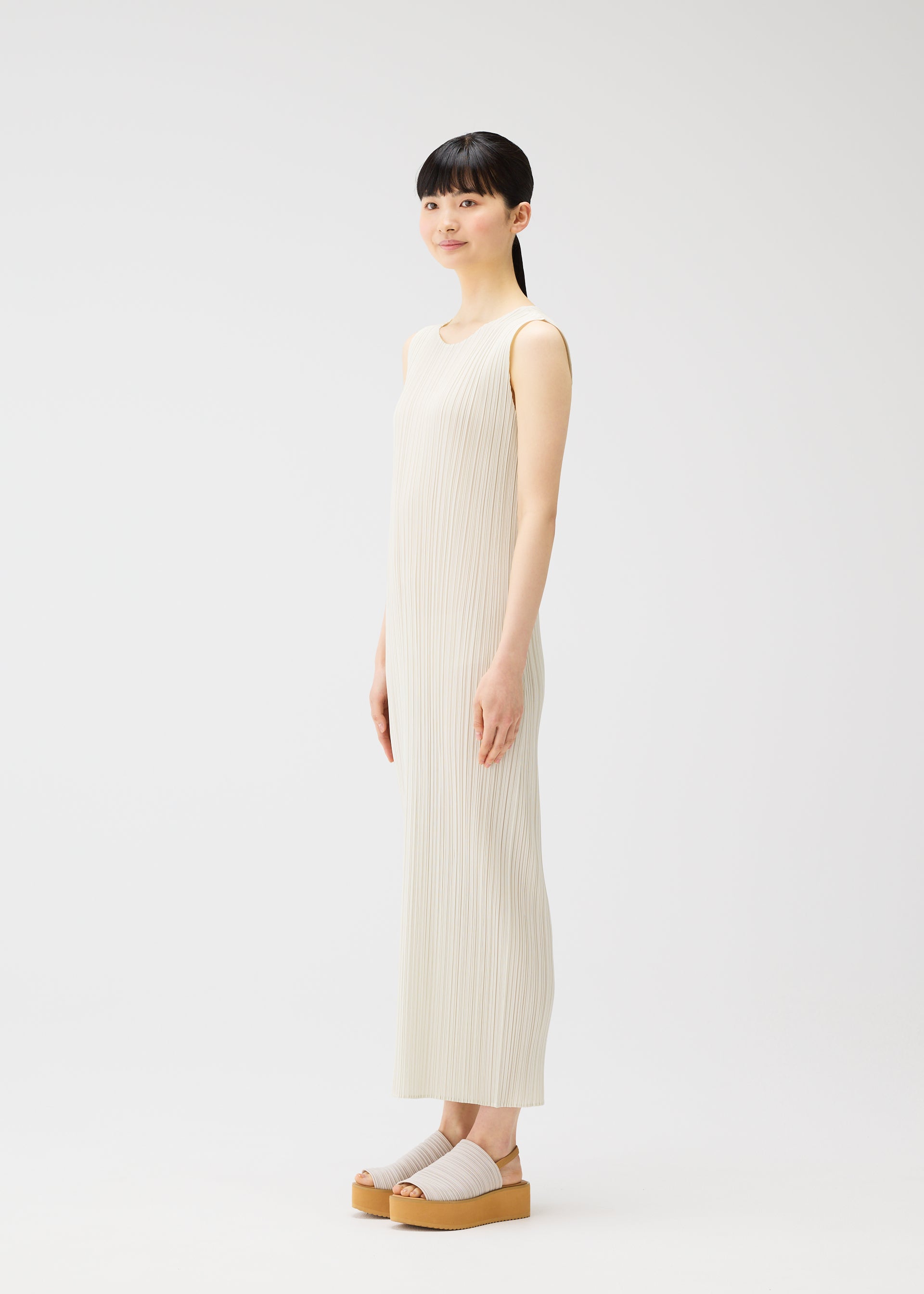 Zoomed view A woman with straight black hair and bangs stands sideways in a long, pleated dress from the MONTHLY COLORS : MAY collection by PLEATS PLEASE ISSEY MIYAKE, paired with beige platform sandals against a plain white background.