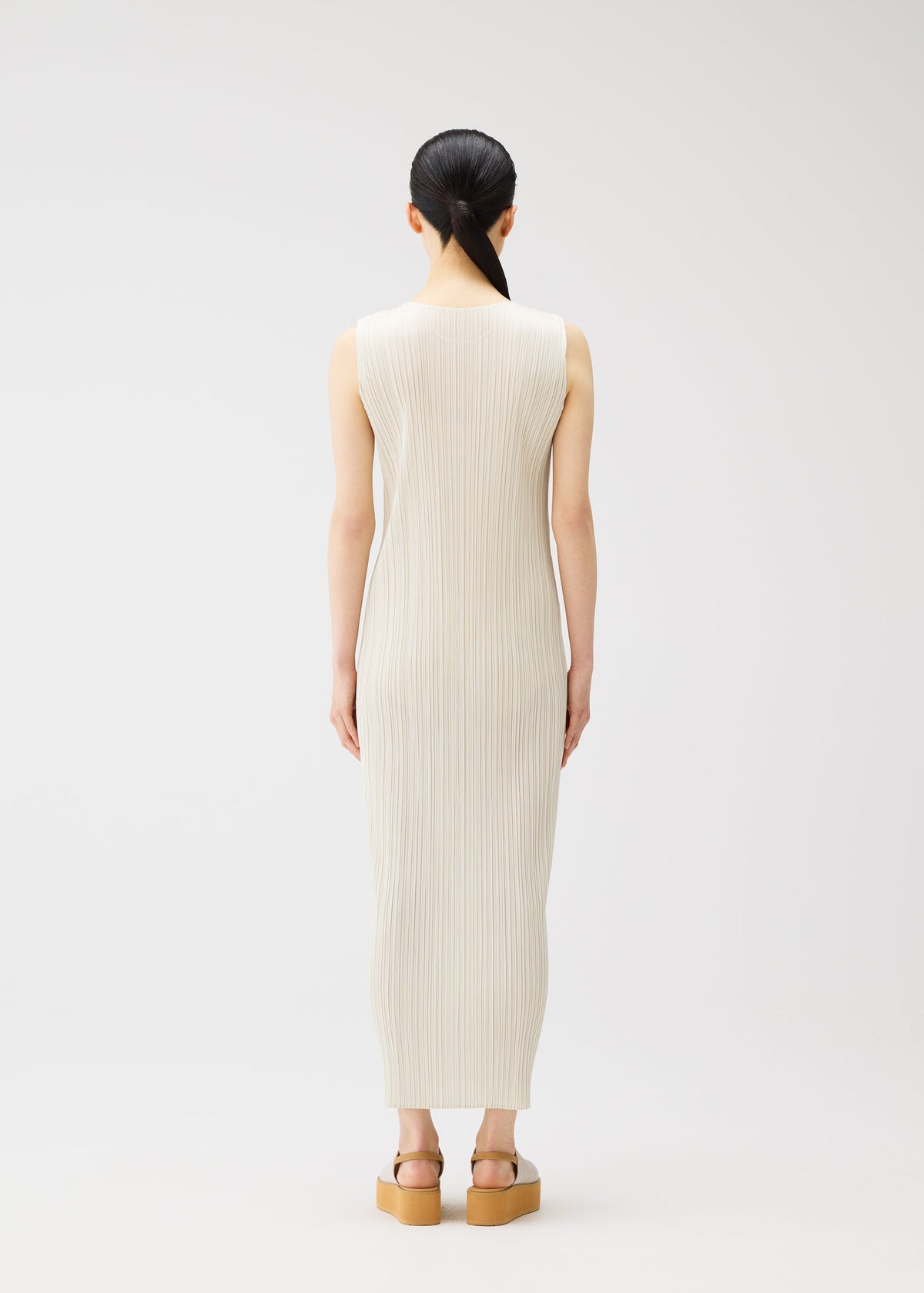 A person with long dark hair in a low ponytail faces away, wearing the sleeveless, ankle-length off-white MONTHLY COLORS : MAY dress by PLEATS PLEASE ISSEY MIYAKE and tan platform sandals against a plain light background. view 9 of 9