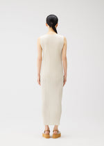 A person with long dark hair in a low ponytail faces away, wearing the sleeveless, ankle-length off-white MONTHLY COLORS : MAY dress by PLEATS PLEASE ISSEY MIYAKE and tan platform sandals against a plain light background.