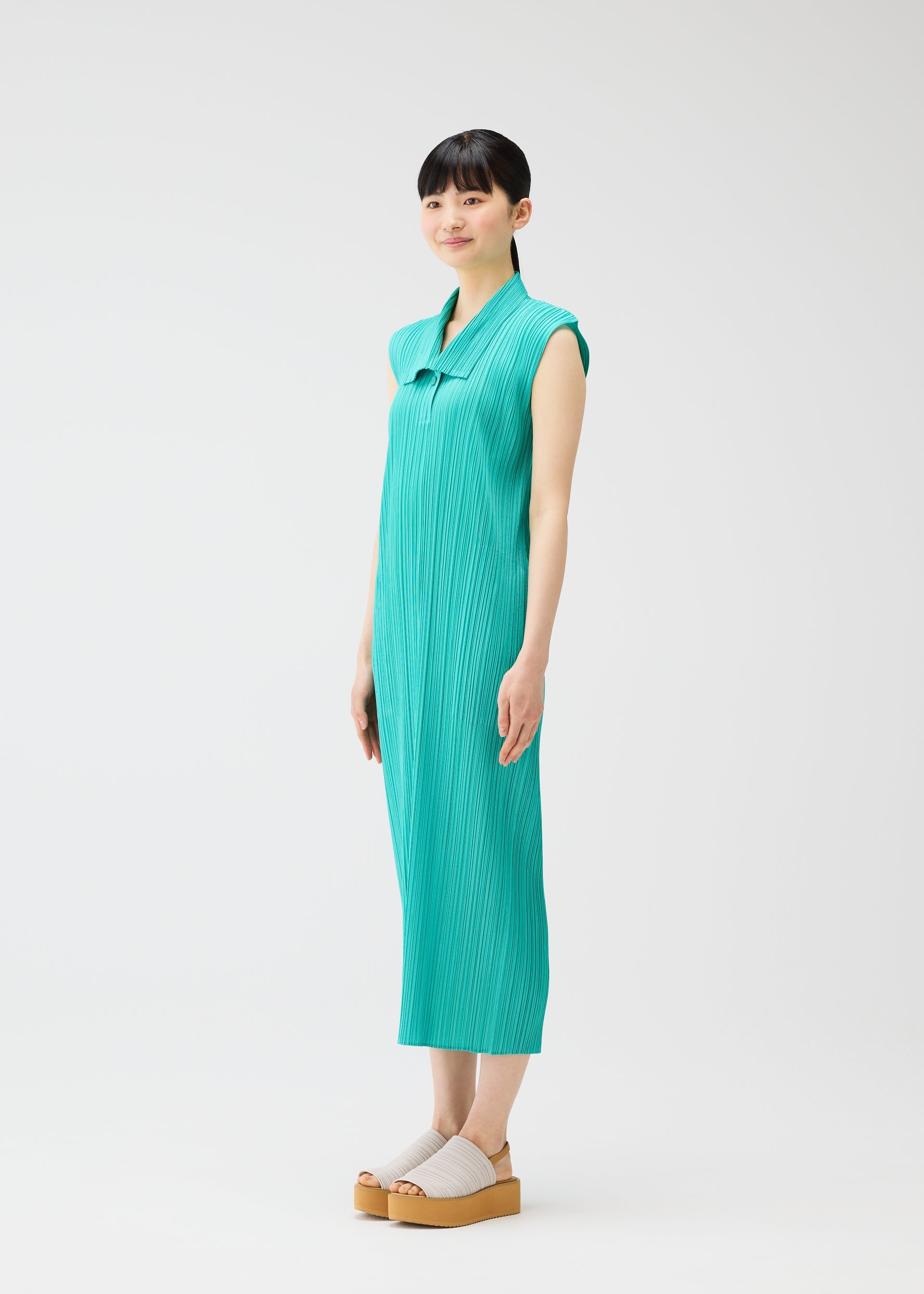 Zoomed view A woman with short dark hair wears the PLEATS PLEASE ISSEY MIYAKE MONTHLY COLORS : MAY sleeveless teal pleated summer dress, styled with white platform sandals, against a plain white background.