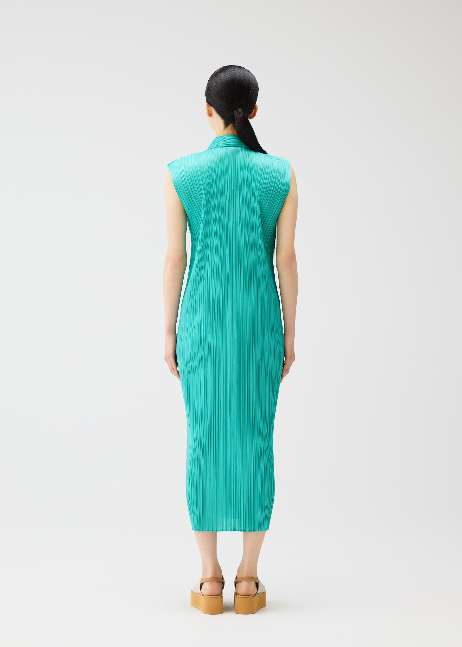 Zoomed view A person with long dark hair in a low ponytail stands facing away, wearing a sleeveless teal pleated dress from PLEATS PLEASE ISSEY MIYAKE's MONTHLY COLORS: MAY and tan platform sandals. The background is plain white.