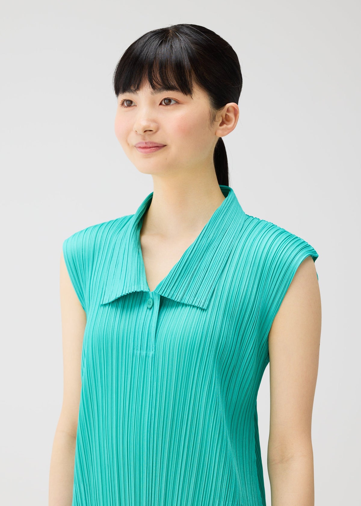 A young woman with straight black hair in a ponytail wears the PLEATS PLEASE ISSEY MIYAKE MONTHLY COLORS : MAY sleeveless dress with a pleated teal top, standing against a plain light background and smiling gently to the side. view 10 of 10
