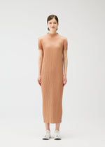 A woman stands facing forward in the PLEATS PLEASE ISSEY MIYAKE MONTHLY COLORS : JUNE light brown, pleated, short-sleeved dress with white sneakers against a plain white background, evoking relaxed coastal hues.