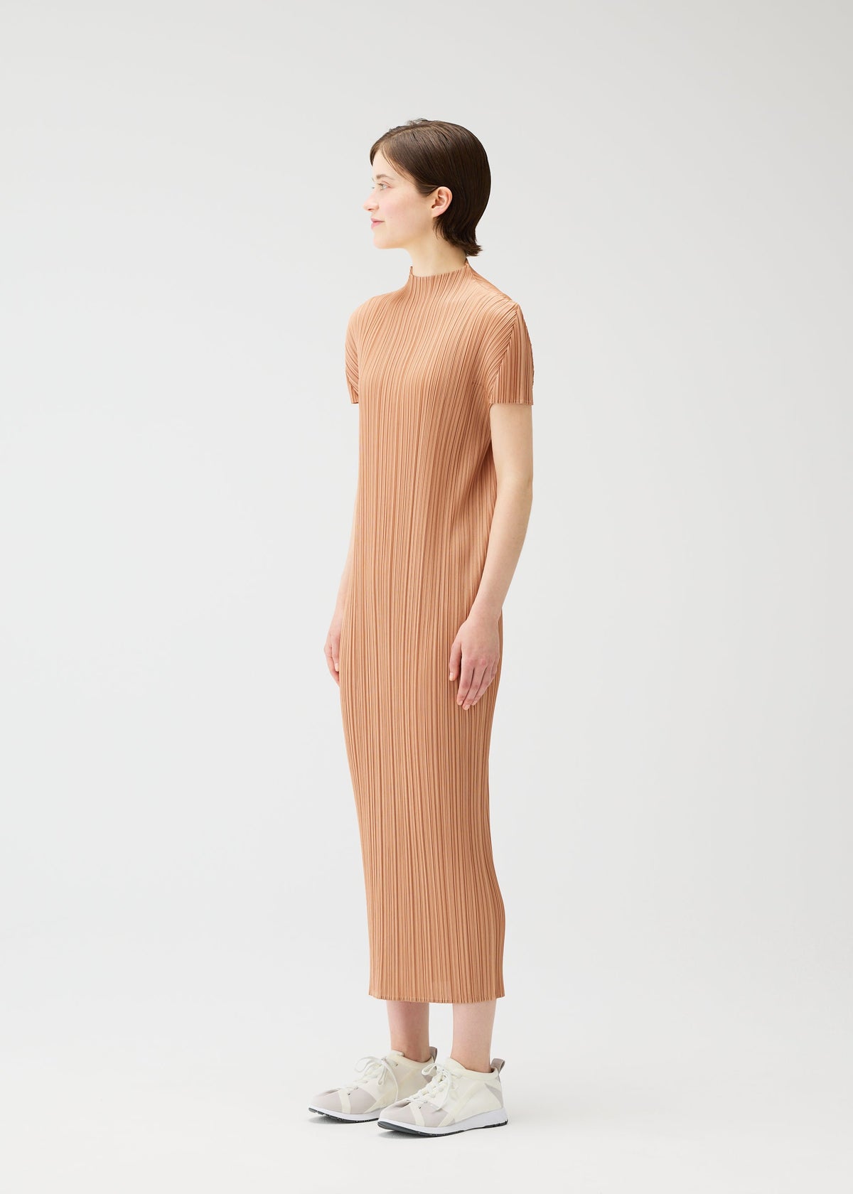 A woman in profile stands against a plain white background wearing the MONTHLY COLORS : JUNE light brown dress by PLEATS PLEASE ISSEY MIYAKE, paired with white sneakers. Her arms rest at her sides as she gazes forward. view 8 of 9
