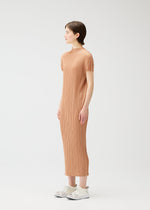 A woman in profile stands against a plain white background wearing the MONTHLY COLORS : JUNE light brown dress by PLEATS PLEASE ISSEY MIYAKE, paired with white sneakers. Her arms rest at her sides as she gazes forward.