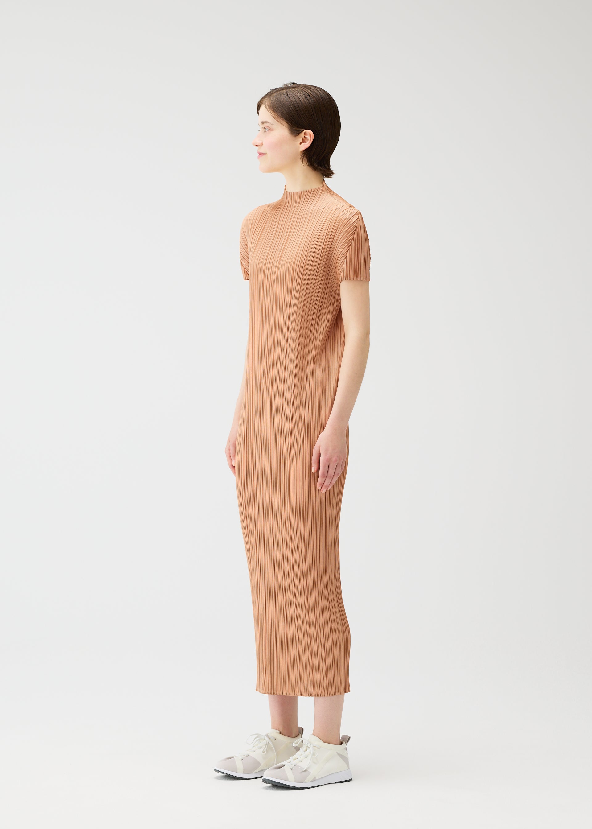 Zoomed view A woman in profile stands against a plain white background wearing the MONTHLY COLORS : JUNE light brown dress by PLEATS PLEASE ISSEY MIYAKE, paired with white sneakers. Her arms rest at her sides as she gazes forward.