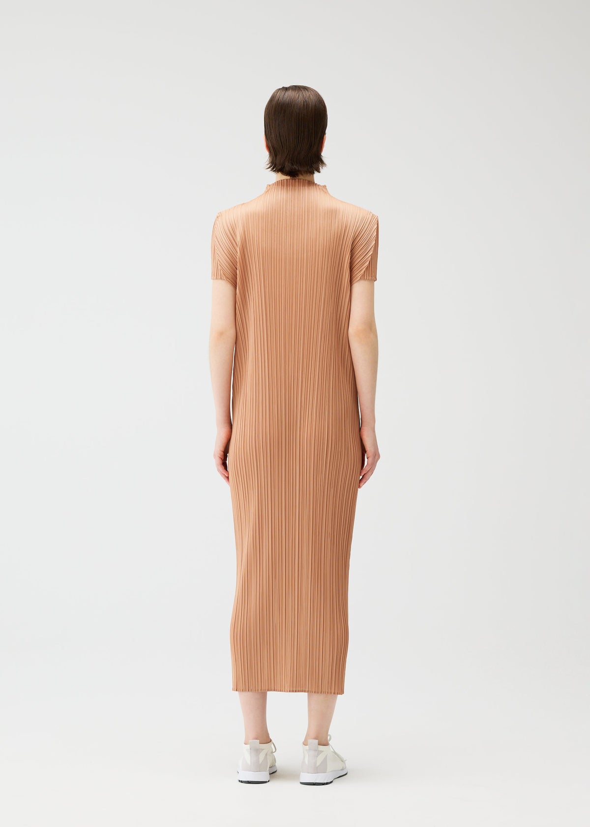 A person stands facing away, wearing the MONTHLY COLORS : JUNE short-sleeved, calf-length ribbed tan high-neck dress by PLEATS PLEASE ISSEY MIYAKE with white sneakers against a plain white background. view 9 of 9