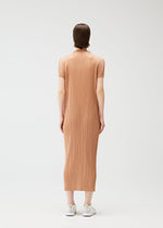 A person stands facing away, wearing the MONTHLY COLORS : JUNE short-sleeved, calf-length ribbed tan high-neck dress by PLEATS PLEASE ISSEY MIYAKE with white sneakers against a plain white background.