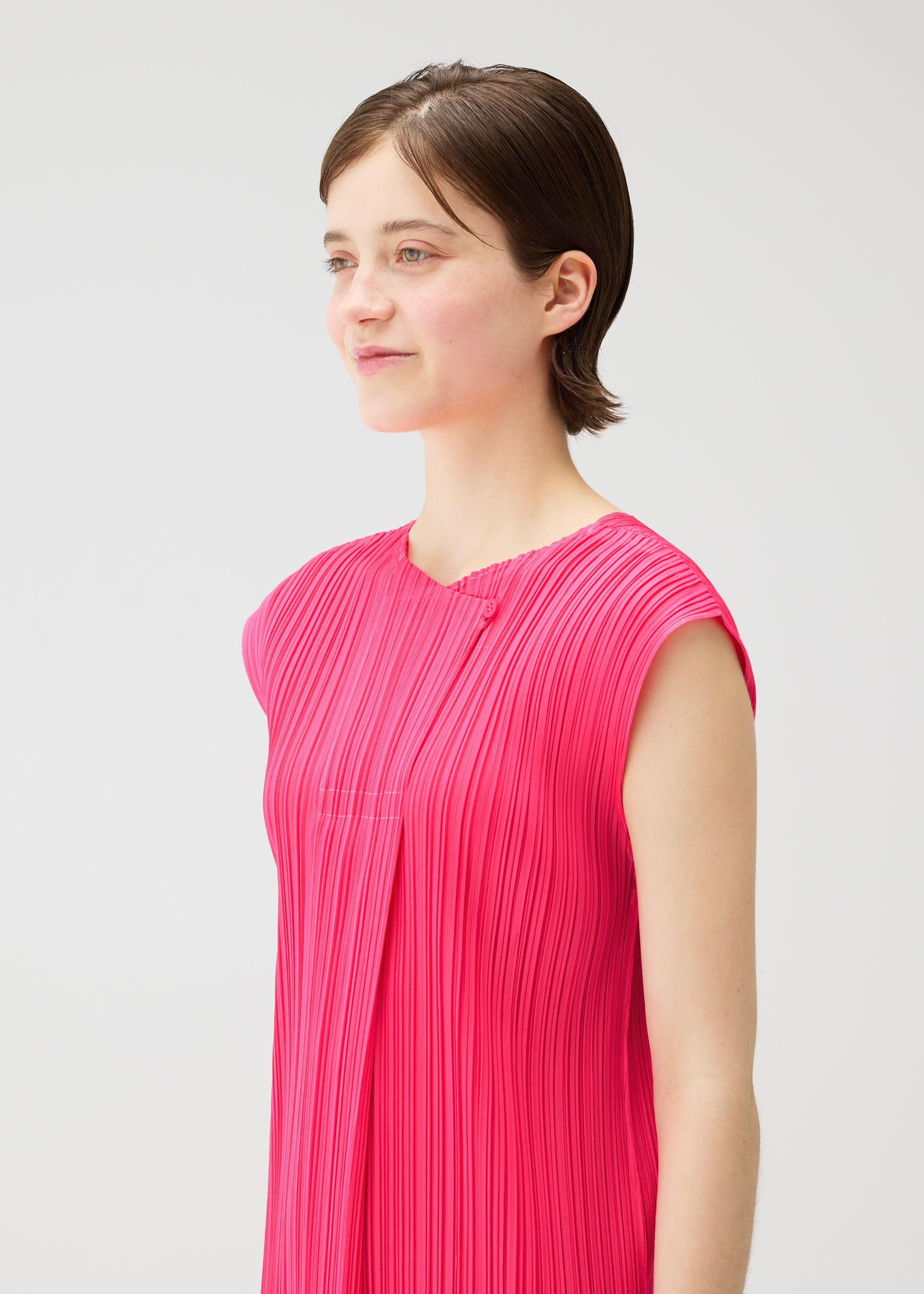 Zoomed view A person with short brown hair models the bright pink, pleated MONTHLY COLORS : JUNE dress by PLEATS PLEASE ISSEY MIYAKE, featuring a sleeveless design and round neck, standing against a plain light background and smiling gently.