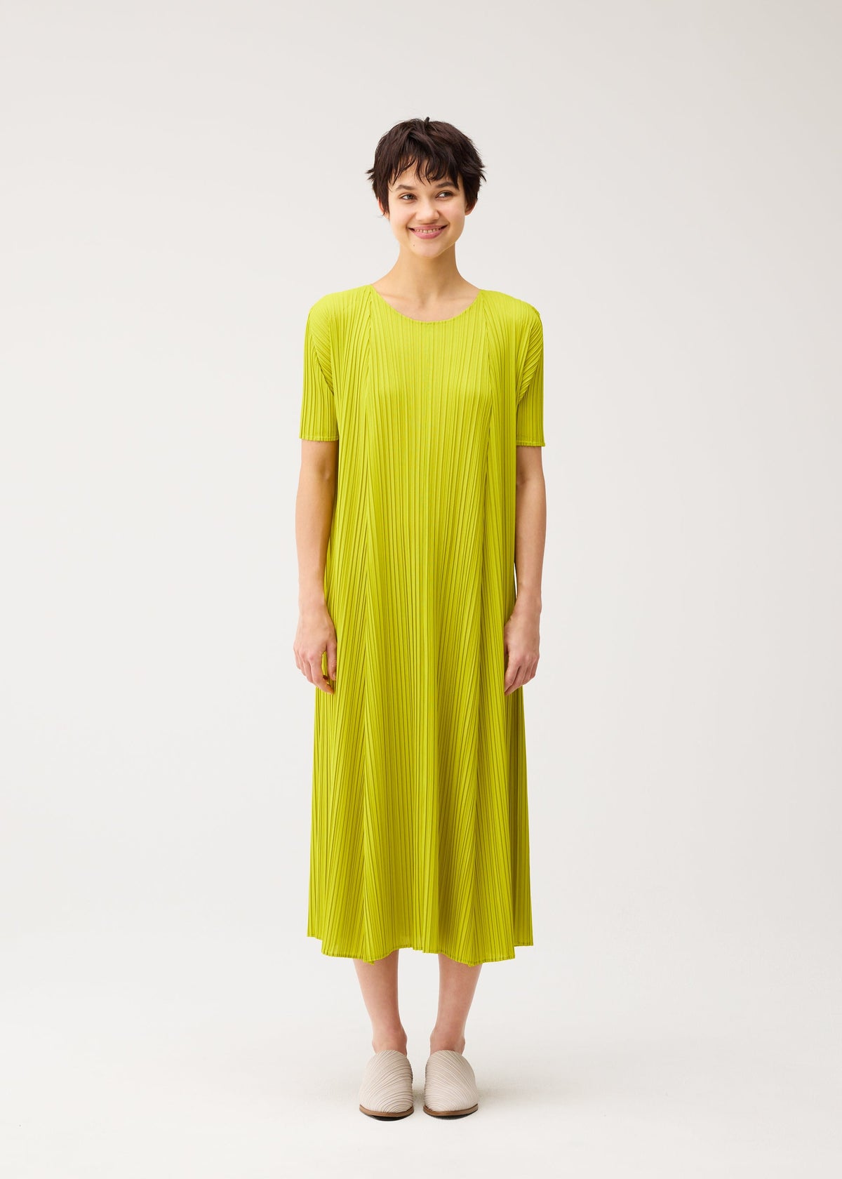 A person with short dark hair stands against a white background, smiling softly in PLEATS PLEASE ISSEY MIYAKE's MELLOW PLEATS—a loose, round-neck yellow-green pleated dress with short sleeves—paired with white slip-on shoes. view 5 of 8