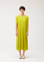 A person with short dark hair stands against a white background, smiling softly in PLEATS PLEASE ISSEY MIYAKE's MELLOW PLEATS—a loose, round-neck yellow-green pleated dress with short sleeves—paired with white slip-on shoes.