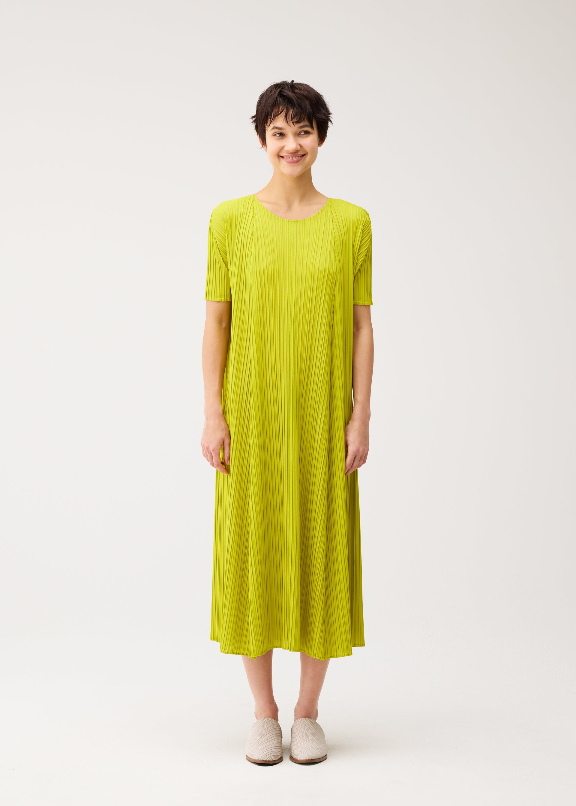 Zoomed view A person with short dark hair stands against a white background, smiling softly in PLEATS PLEASE ISSEY MIYAKE's MELLOW PLEATS—a loose, round-neck yellow-green pleated dress with short sleeves—paired with white slip-on shoes.