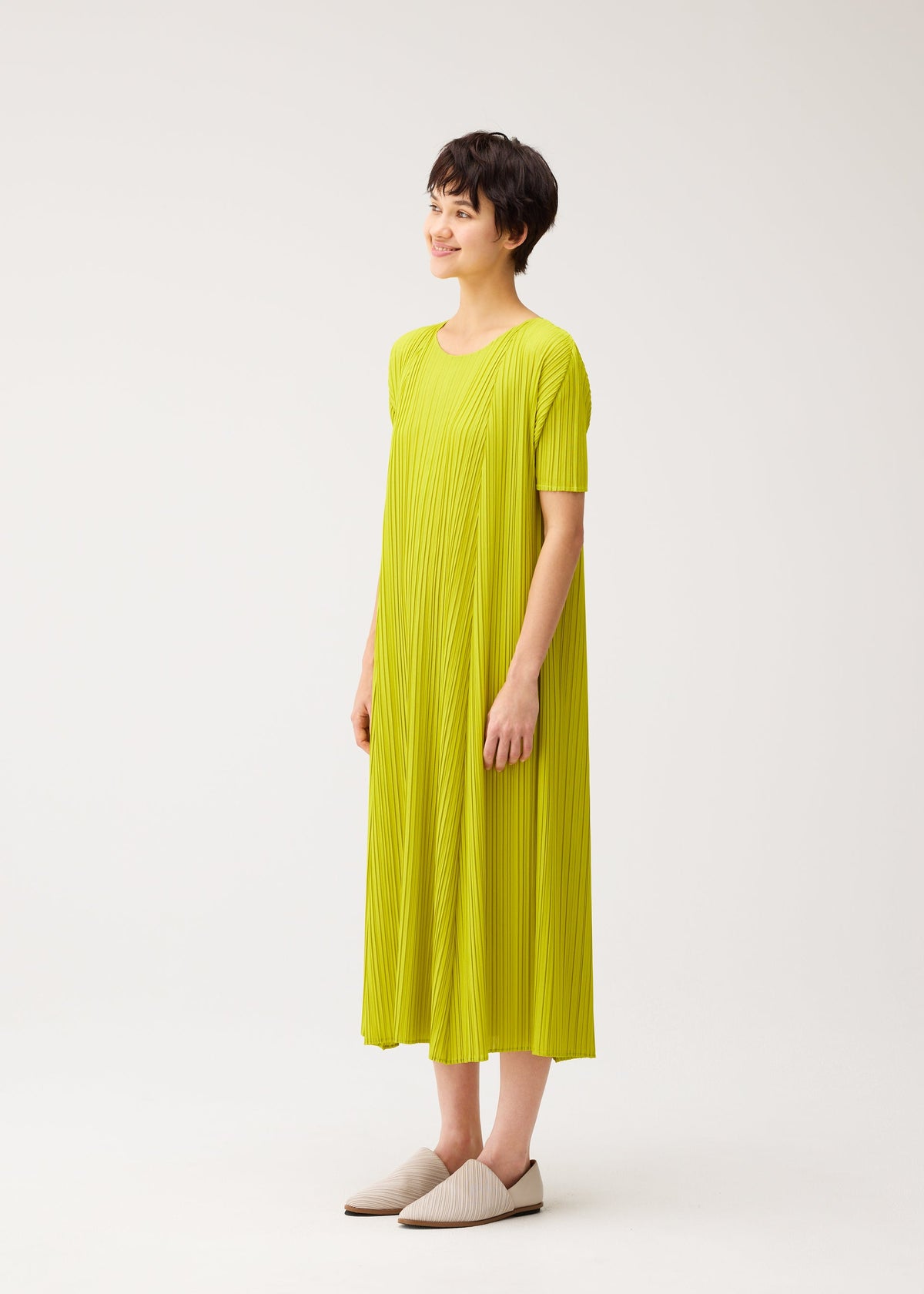 A person with short dark hair smiles in a loose-fitting, bright yellow-green MELLOW PLEATS dress from PLEATS PLEASE ISSEY MIYAKE and light slip-on shoes, against a plain white background. view 6 of 8
