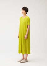 A person with short dark hair smiles in a loose-fitting, bright yellow-green MELLOW PLEATS dress from PLEATS PLEASE ISSEY MIYAKE and light slip-on shoes, against a plain white background.