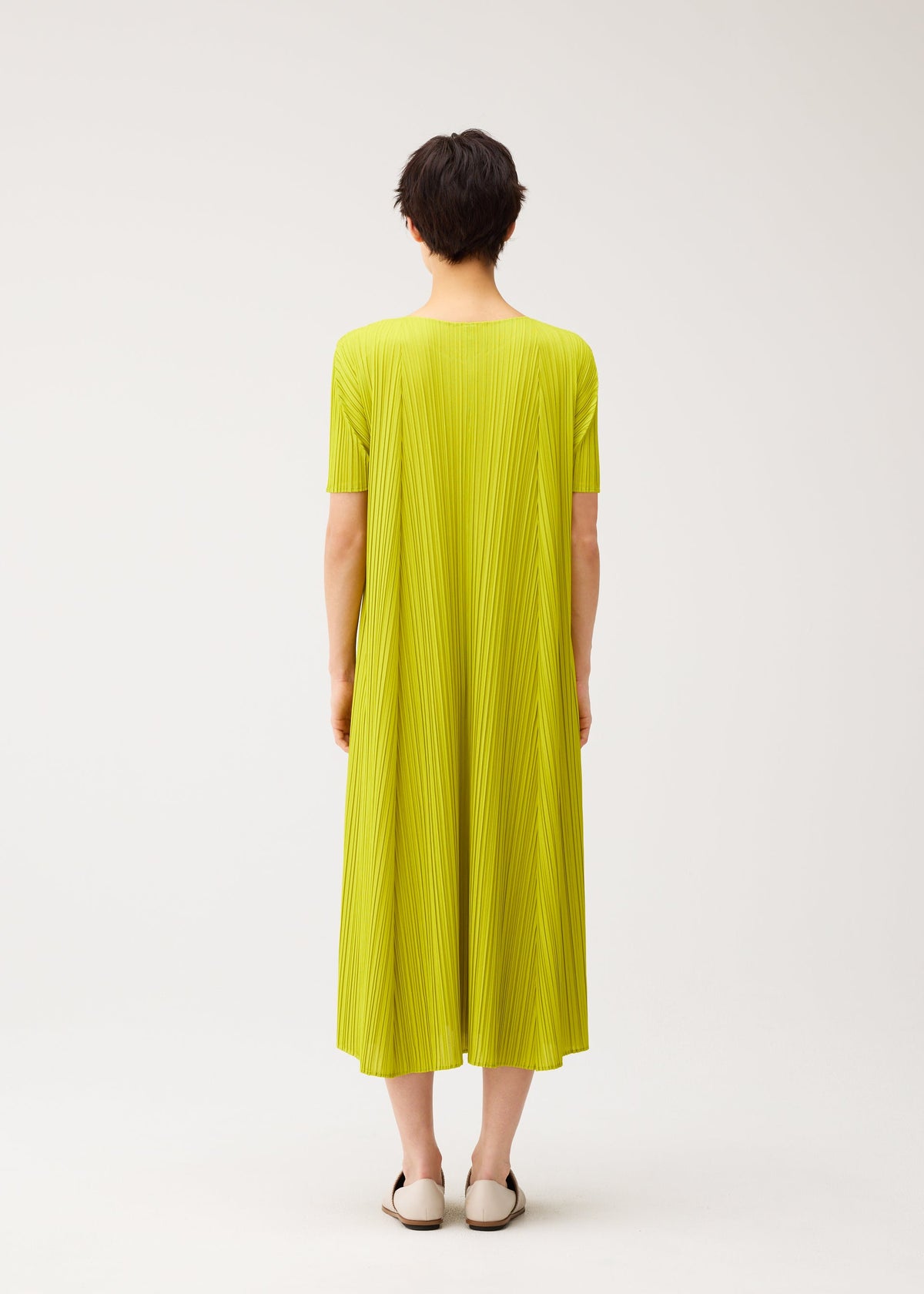 A person with short dark hair faces away, wearing the MELLOW PLEATS dress by PLEATS PLEASE ISSEY MIYAKE in bright yellow-green with a round neck and pleated short sleeves, paired with white slip-on shoes against a plain background. view 7 of 8
