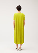 A person with short dark hair faces away, wearing the MELLOW PLEATS dress by PLEATS PLEASE ISSEY MIYAKE in bright yellow-green with a round neck and pleated short sleeves, paired with white slip-on shoes against a plain background.